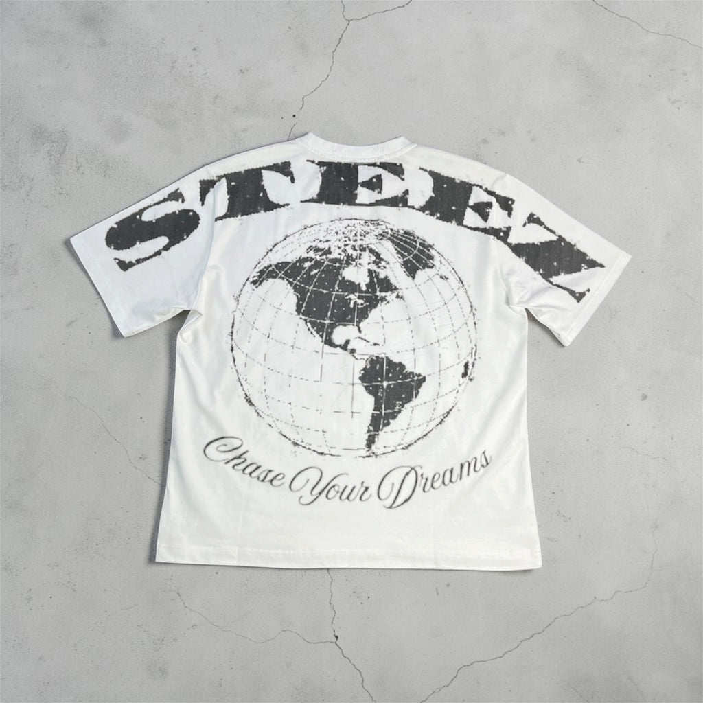 A white short-sleeve graphic T-shirt laid flat on a textured gray concrete surface. The back of the shirt features a large distressed black 'STEEN' logo arched over a black globe graphic, with 'Chase Your Dreams' written in cursive below.