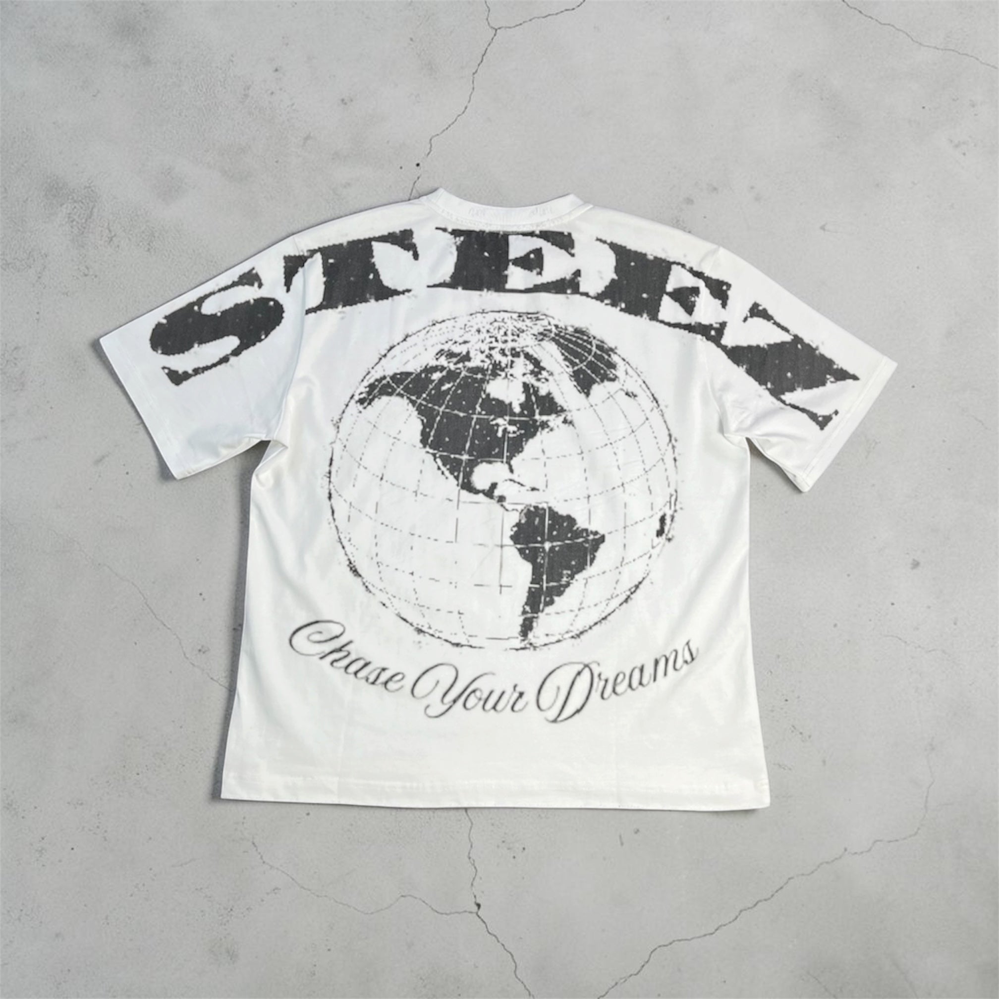 A white short-sleeve graphic T-shirt laid flat on a textured gray concrete surface. The back of the shirt features a large distressed black 'STEEN' logo arched over a black globe graphic, with 'Chase Your Dreams' written in cursive below.