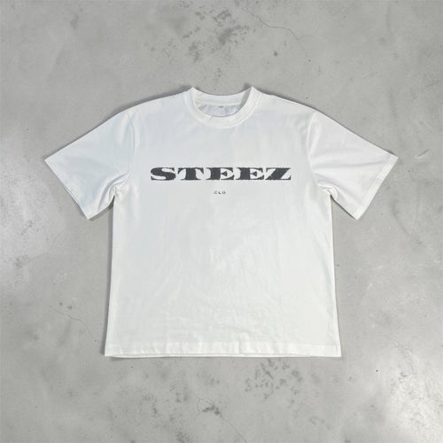 A white short-sleeve crewneck t-shirt with a black 'STEEZ' logo printed across the chest, laid flat on a textured gray concrete surface.